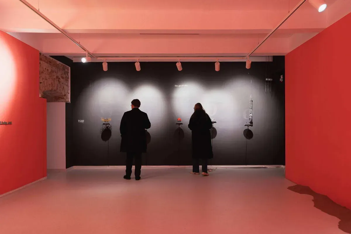 Doi vizitatori într-o sală de expoziție cu pereți roșii, în fața unui perete negru luminat de spoturi