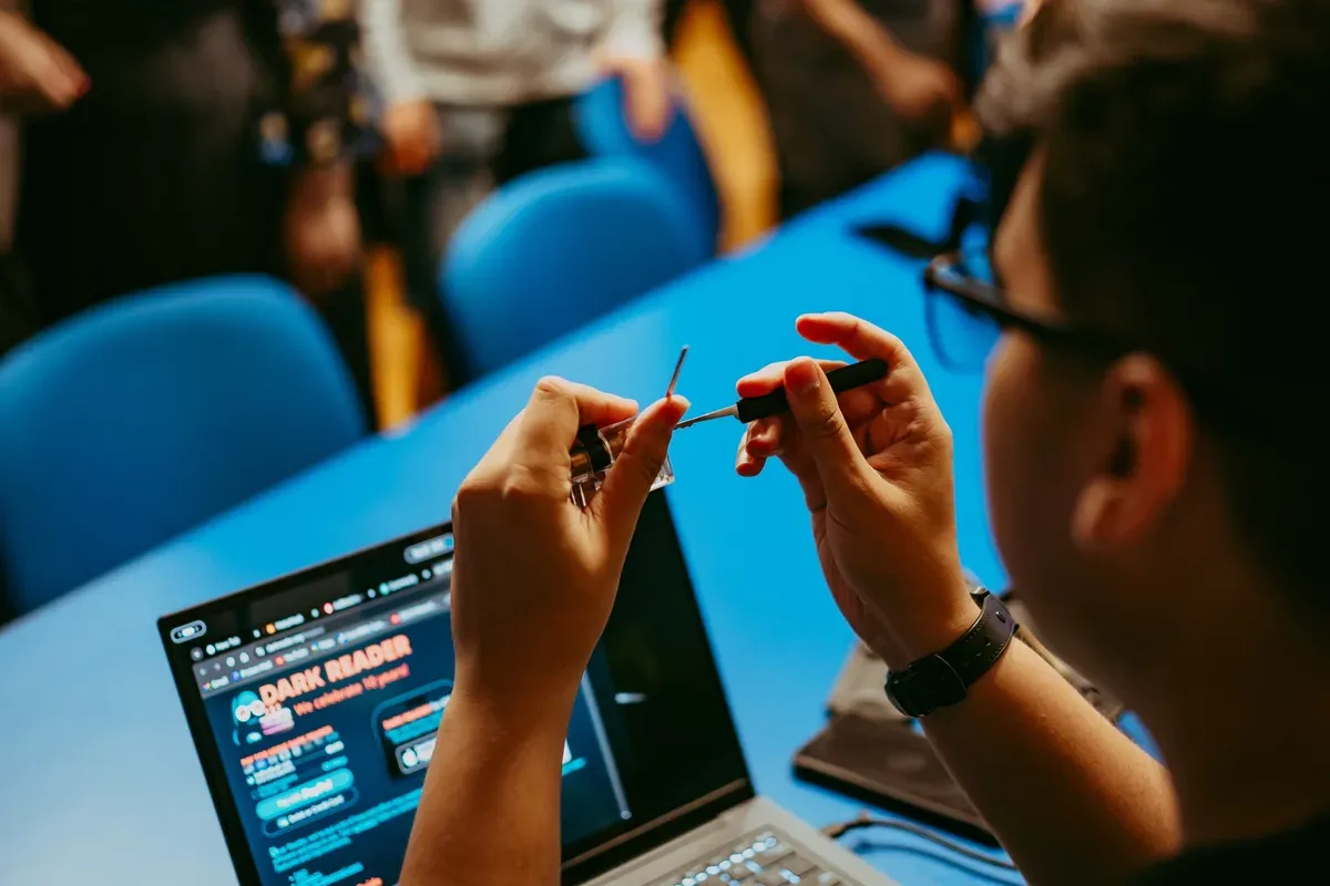 Mâinile unui participant lucrând cu o unealtă fină, lângă un laptop